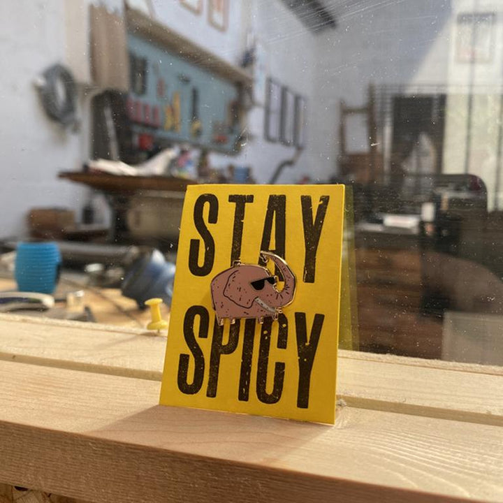 A pink elephant pin on a yellow backing that says stay spicy.